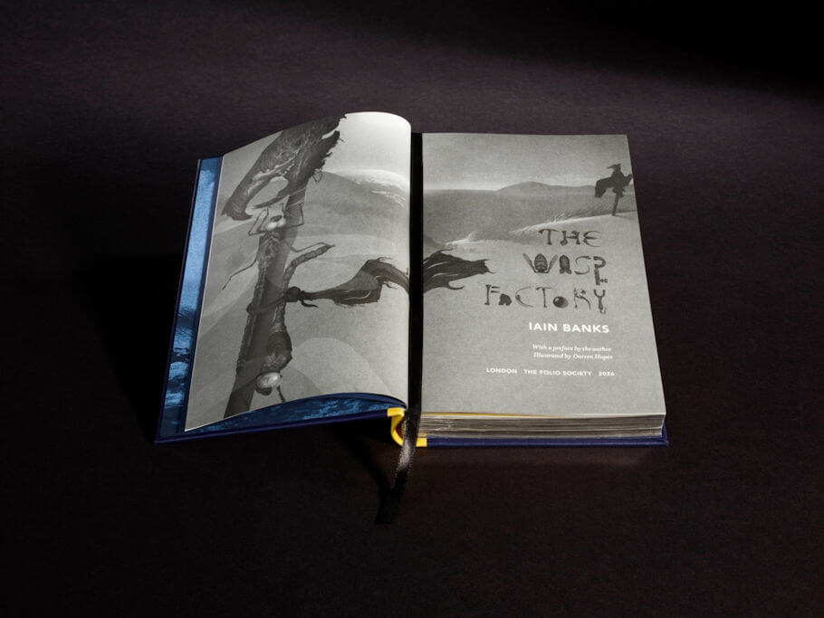 The same book lies open to its title pages on a dark surface, showing grayscale artwork that stretches across both pages. The left page shows a twisted, gnarled post or tree trunk hung with skull-like objects against rolling hills, while the right page continues the landscape with a lone scarecrow-like figure standing on a distant dune.